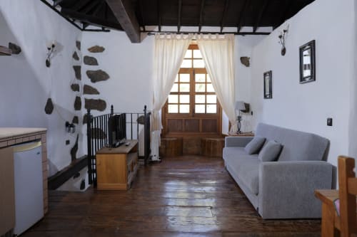 Rustic cottage interior at Casonas de Marengo with wooden beams, white walls, gray sofa, crib, and traditional décor