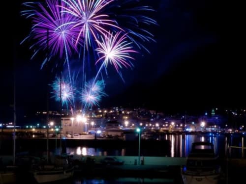 I fuochi d'artificio illuminano il cielo notturno sopra il porto con edifici e barche a Can Ricós, Vallgorguina