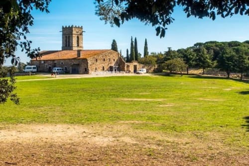 Chiesa in pietra medievale con campanile circondata da prato verde e cipressi presso l'ostello rurale Can Ricós a Vallgorguina