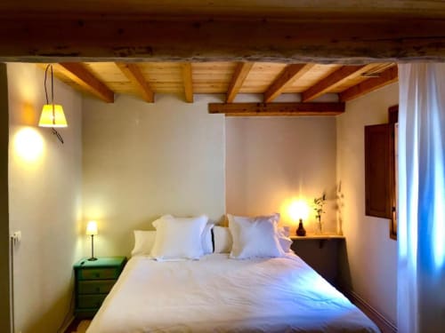 Dormitorio acogedor en la casa rural Casa Tapiolas en Molló con vigas de madera vista, ropa de cama blanca, iluminación cálida y vistas a la montaña