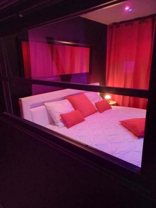 Chambre moderne à La Maison du Plaisir : éclairage rose néon, lit blanc, coussins rouges et ambiance tamisée