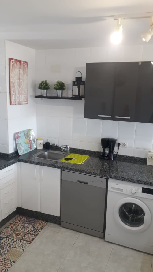 Cocina compacta moderna con gabinetes blancos, encimera negra, fregadero, lavavajillas, lavadora y estante decorativo