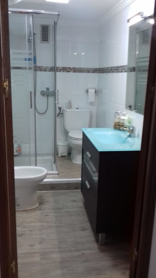 Baño moderno con lavamanos turquesa, ducha con mampara de cristal, inodoro y paredes alicatadas en apartamento de Puerto Santa María