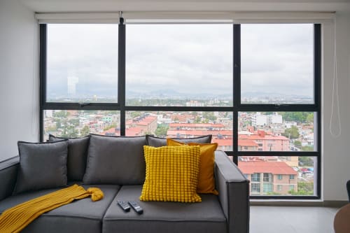 Departamento moderno en CDMX con sofá gris, acentos amarillos y vistas panorámicas de la ciudad desde ventanales