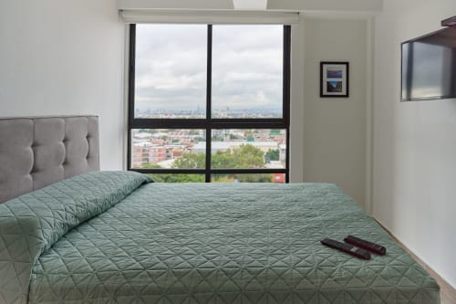 Recámara moderna en la Ciudad de México con cama acolchada verde, cabecera gris y amplia vista del horizonte urbano a través de grandes ventanales
