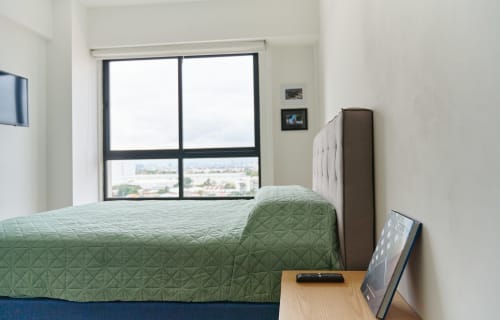 Dormitorio moderno en apartamento de la Ciudad de México con vista de la ciudad a través de grandes ventanales, ropa de cama acolchada verde y decoración minimalista