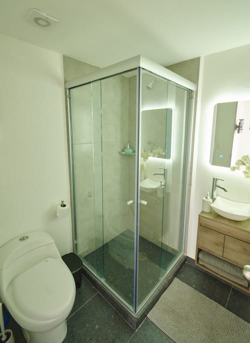 Baño moderno en apartamento Pares Homes CDMX con ducha de cristal, inodoro blanco, espejo iluminado y madera
