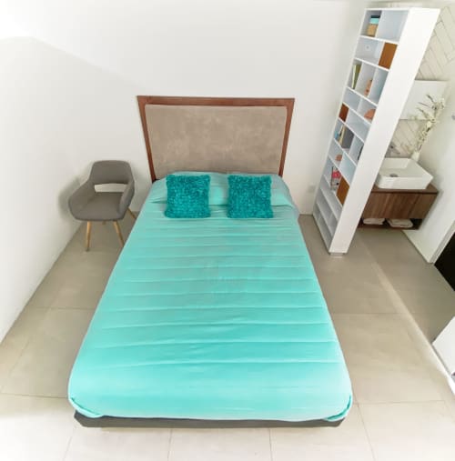 Habitación moderna en Pares Homes CDMX con cama turquesa, cabecera de madera, silla gris y estantería blanca.