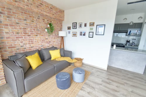 Sala de estar moderna en apartamento de CDMX con ladrillo expuesto, sofá gris, detalles amarillos y área de cocina en Pares Homes