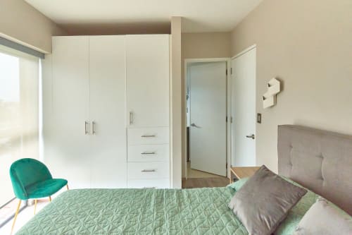 Recámara moderna en CDMX con armario blanco, cama acolchada verde, silla turquesa y vista a la ciudad