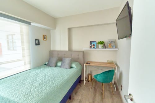 Recámara moderna en apartamento Pares Homes CDMX con ropa de cama verde menta, TV de pared, escritorio con silla verde y vistas a la ciudad.