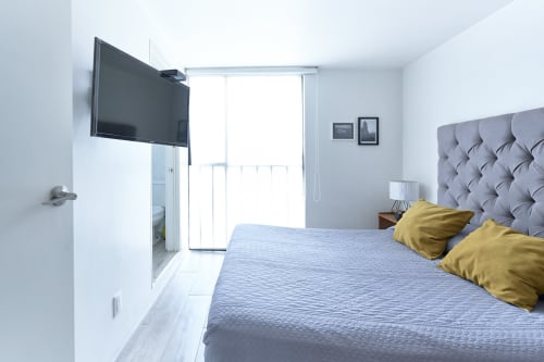 Dormitorio moderno en apartamento Pares Homes, Ciudad de México, con cama tapizada gris, almohadas amarillas, TV de pared y