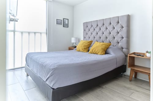 Recámara moderna en apartamento Pares Homes, Ciudad de México, con cabecera capitonada gris, almohadas amarillas y mesita de noche de madera