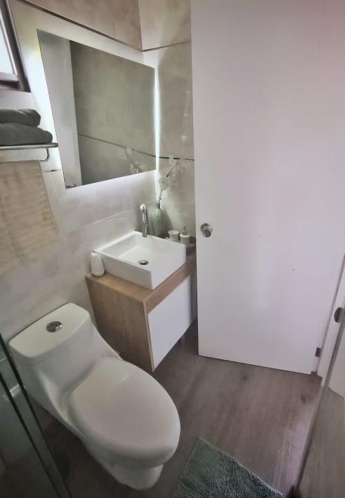 Baño moderno en departamento Pares Homes, CDMX con lavamanos blanco, inodoro, espejo iluminado y área de ducha alicatada