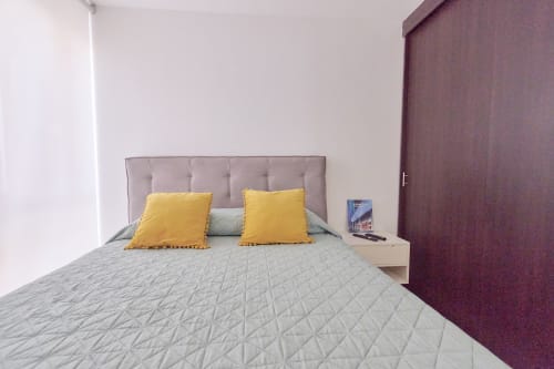 Moderno dormitorio en apartamento Pares Homes CDMX con cama acolchada gris, cabecera taupes, almohadas amarillas y burdeos