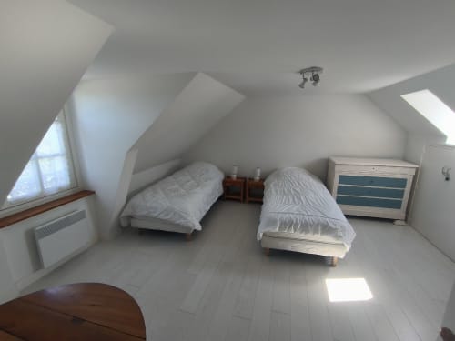 Chambre mansardée lumineuse avec deux lits simples, draps blancs, tables de nuit en bois, commode et fenêtres de toit