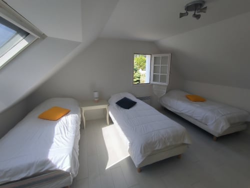 Chambre lumineuse sous les toits avec trois lits simples, oreillers orange, fenêtre ouverte sur le jardin du gîte Locmaria, Belle Île en