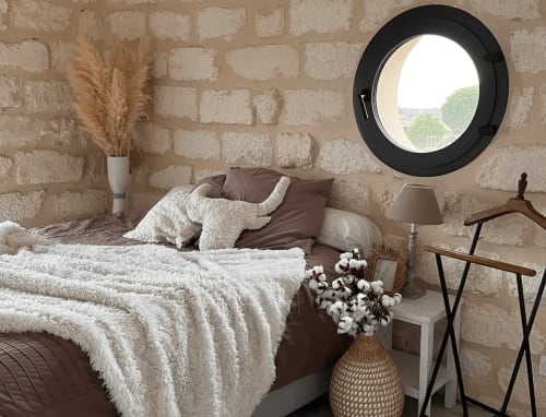 Chambre douillette à ADELOUANE aux murs de pierre, fenêtre circulaire, literie crème et décor rustique à Aigues-mortes