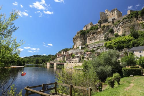 Medieval castle perched on cliff above river in Carsac-Aillac, France, with canoes on water and green countryside