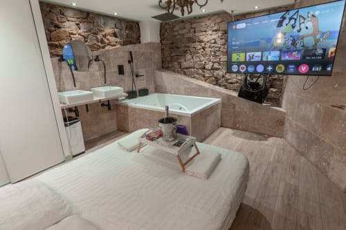Découvrez la Love Room 3, une suite romantique idéale pour une escapade en couple. Avec ses magnifiques murs en pierre apparents, cette chambre offre une ambiance chaleureuse et authentique. Profitez d'une double vasque moderne et d'un grand écran TV avec Netflix pour vos soirées détente. Le clou du séjour ? Une superbe baignoire à balnéothérapie pour deux, parfaite pour un moment de relaxation absolue. Réservez la Love Room 3 et vivez une expérience inoubliable.