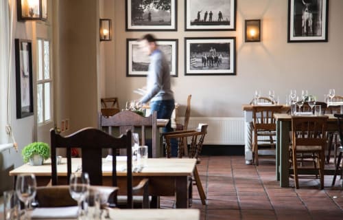 Three Little Pigs restaurant dining room with wooden tables, framed black and white artwork, and server in motion at