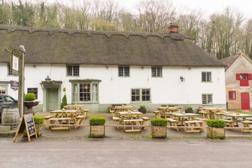 Three Little Pigs pub exterior with thatched roof, white walls, and wooden picnic tables in beer garden, Blandford Forum