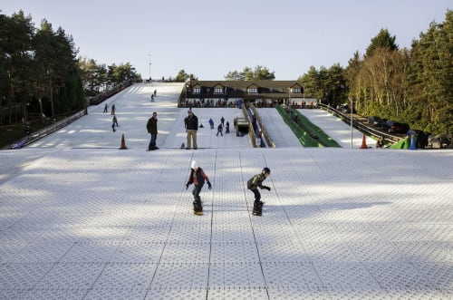 Skiers on indoor ski slope at Three Little Pigs hotel in Blandford Forum with lodge building and forest backdrop