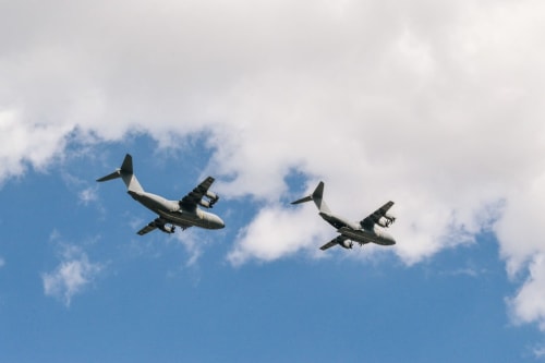 Two military transport aircraft flying in formation against a cloudy blue sky