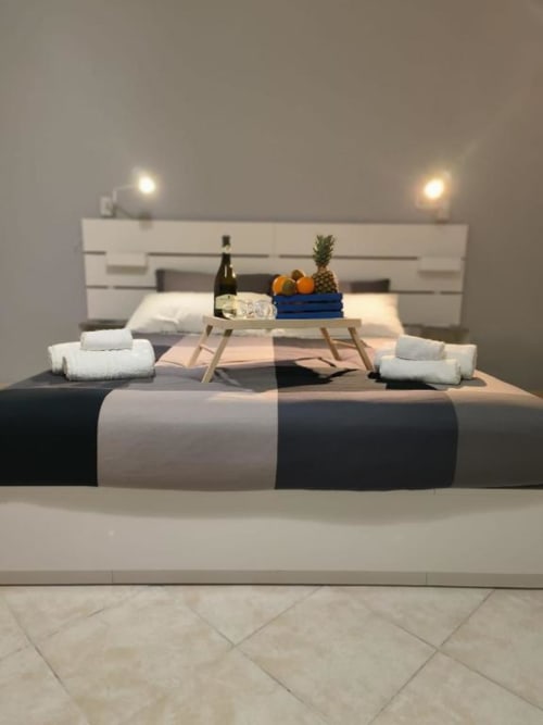 Camera moderna a Sikelia House Cefalù con letto a righe, testiera bianca, vassoio in legno con vino e frutta, asciugamani arrotolati