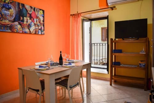 Zona pranzo dell'appartamento Sikelia House a Cefalù con pareti arancioni, tavolo in legno, sedie e balcone con vista sulla città