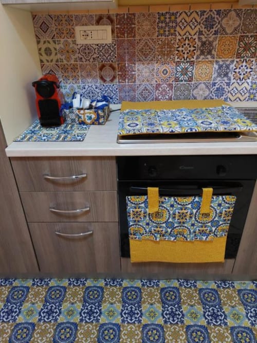 Zona cucina di Sikelia House Cefalù con piastrelle decorate, fornello, asciugamano giallo e blu, caffettiera e