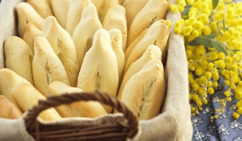 The *navettes de Marseille*, traditional boat-shaped biscuits, are neatly arranged in a basket. Their golden, slightly cracked texture reflects their perfect baking. Beside the basket, a bouquet of yellow flowers adds a bright, natural touch. These iconic biscuits are often associated with Candlemas and have a light orange blossom flavor, a typical characteristic of the Marseille recipe.