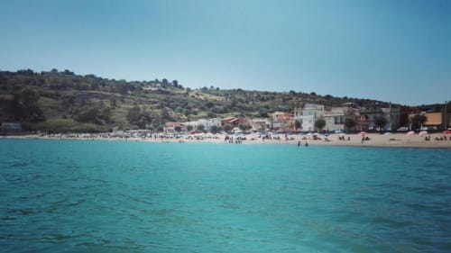 Mare Mediterraneo turchese con spiaggia sabbiosa, edifici colorati e il villaggio collinare di Agnone Bagni, Italia