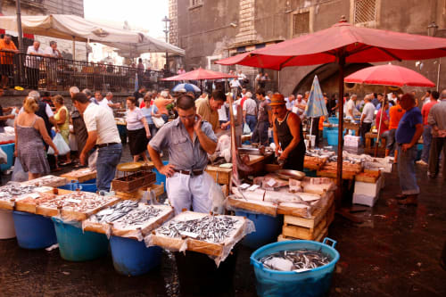 Bustling Mediterranean fish market in Agnone Bagni with vendors, red umbrellas, and fresh seafood displays