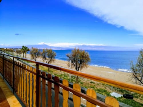 Wooden balcony railing overlooking sandy beach, blue sea, palm trees, and snow-capped Mount Etna at Noemi Holiday House,