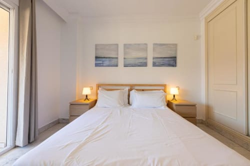 Bright modern bedroom at Marbella Home Rentals with double bed, wooden nightstands, bedside lamps, and coastal artwork in
