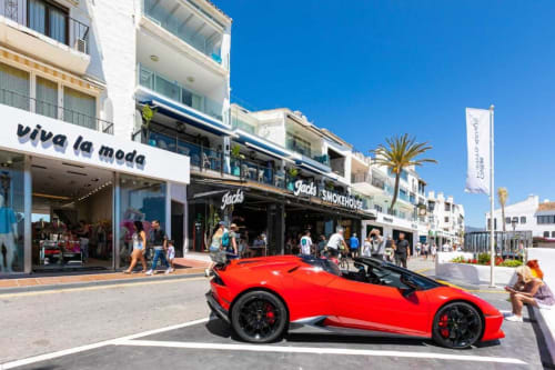 Red sports car parked at Puerto Banús Marina, Marbella with shops and beachfront buildings in background