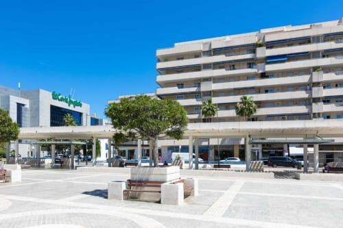 Modern apartment building in Puerto Banús, Marbella with ground floor shops and wide plaza featuring trees and seating areas