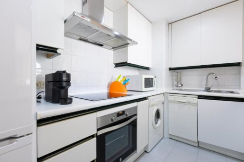 Modern white kitchen with black trim, stainless appliances, and orange knife block in Marina Banús apartment