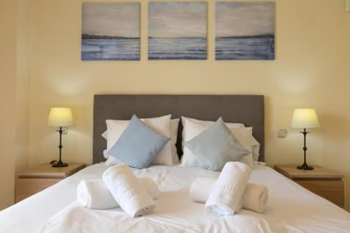 Elegant bedroom at Marbella Home Rentals with gray headboard, white bedding, blue pillows, and coastal artwork above in
