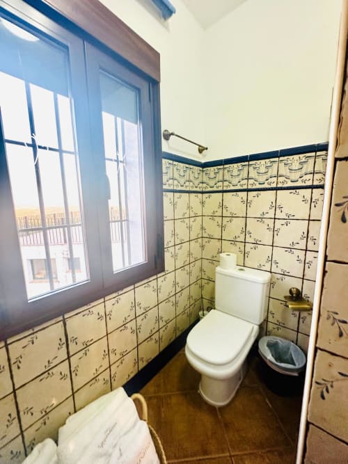 Baño con paredes de azulejos decorativos, inodoro blanco, piso de madera y amplia ventana con vista a Mérida
