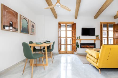 Comedor con sillas verdes, sofá amarillo y chimenea eléctrica en salón luminoso de apartamento turístico en Granada.