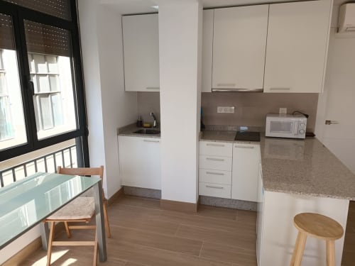Cocina moderna con muebles blancos, barra de granito y mesa con silla junto a ventana en apartamento de Granada.