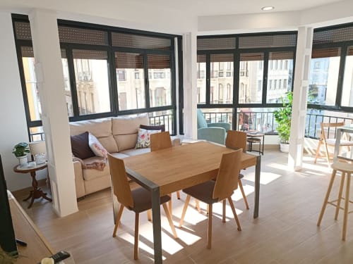 Sala luminosa con comedor de madera, sofá, sillón y amplios ventanales con vistas urbanas en apartamento de Granada.