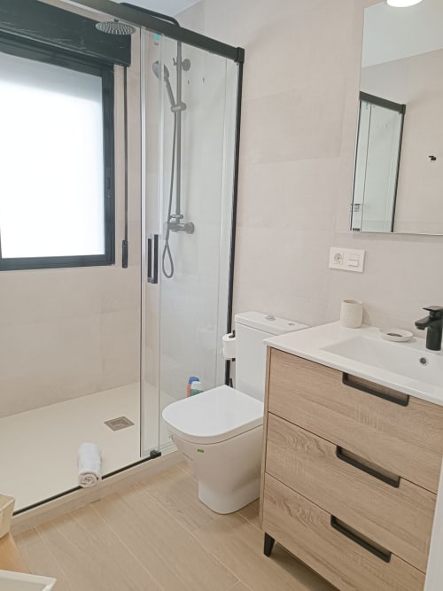 Baño moderno con ducha de vidrio, lavabo con mueble de madera y ventana luminosa en apartamento turístico en Granada.