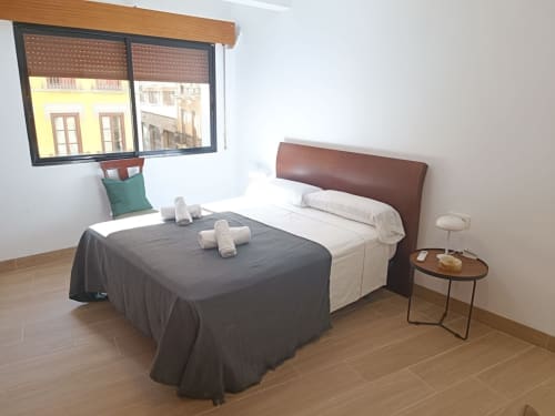 Dormitorio luminoso con cama doble, cabecero marrón y ventana con vistas a la ciudad en apartamento cerca de Plaza Nueva.