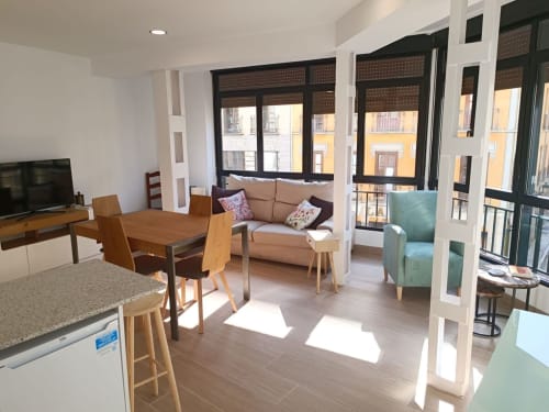 Sala de estar luminosa con sofá, sillón, comedor de madera y grandes ventanales en apartamento céntrico de Granada.