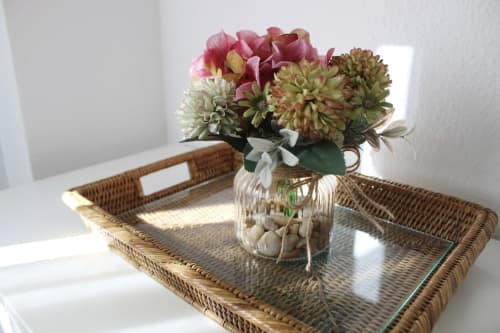 Detalle decorativo con flores artificiales en un jarrón de cristal sobre una bandeja de mimbre en apartamento turístico.