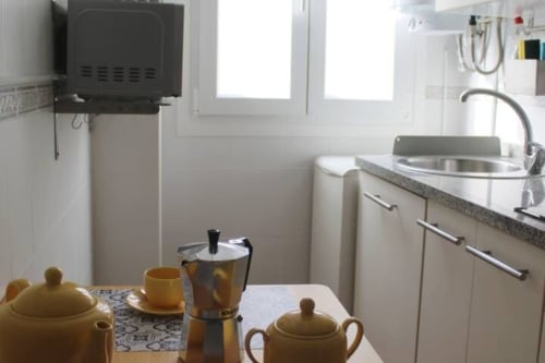 Cocina compacta con mesa, cafetera italiana y vajilla amarilla junto a fregadero y ventana en apartamento de Granada.