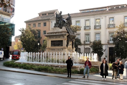 Fuente y monumento a Isabel la Católica y Cristóbal Colón en una plaza céntrica de Granada, rodeados de visitantes.